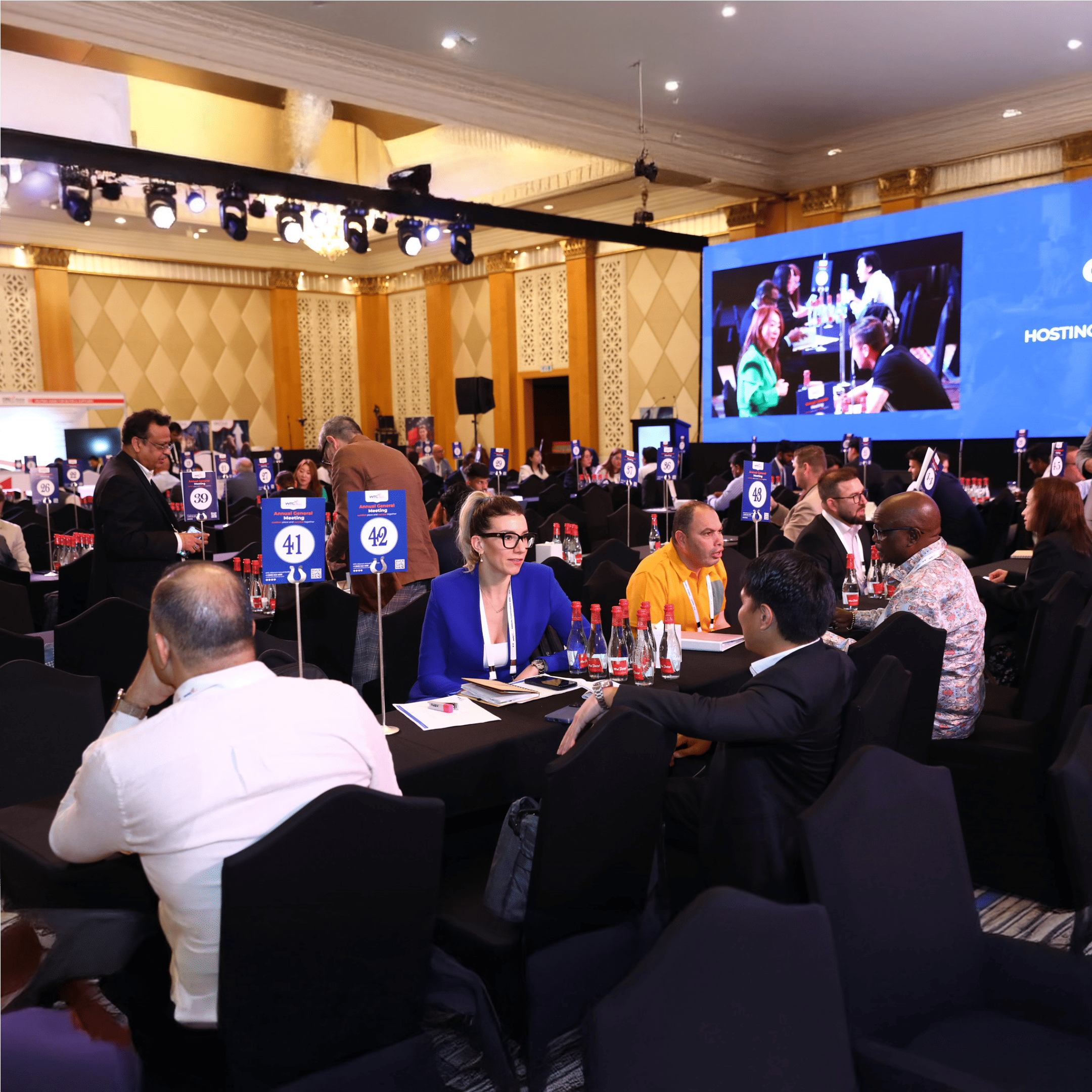  Large conference room filled with groups of professionals having networking discussions at numbered tables, with water bottles and notebooks on the tables, and a presentation screen displaying meeting activity in the background.